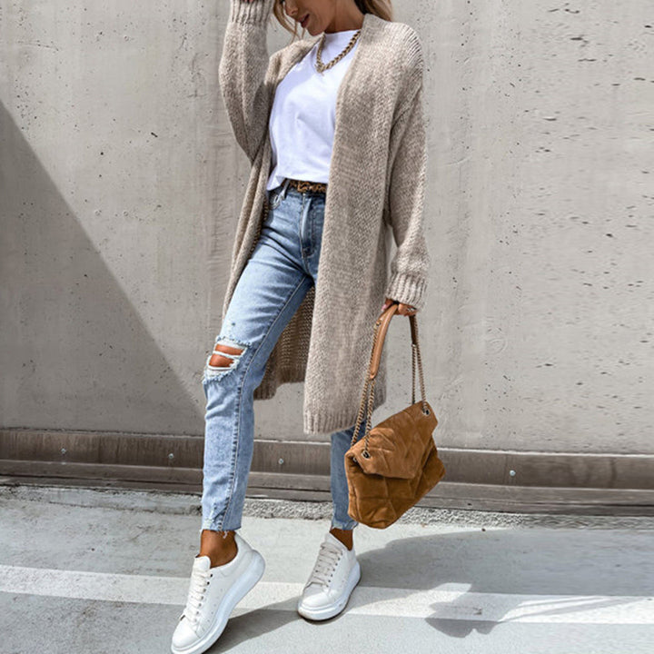 Frau in lässigem Outfit mit langem Strickcardigan, weißem T-Shirt, zerrissenen Jeans und weißen Sneakers, hält braune Handtasche vor Betonwand.