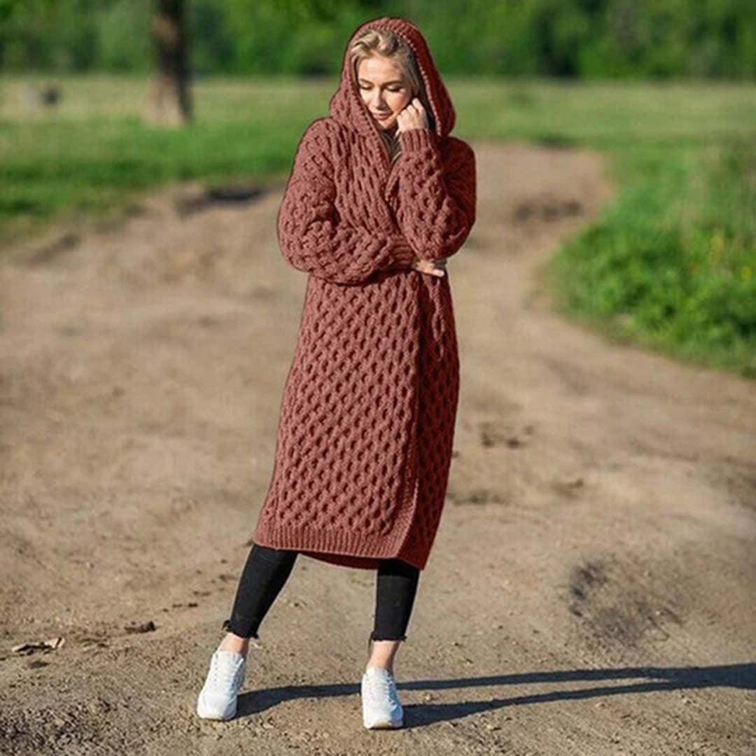 Frau in langem, braunem Strickmantel mit Kapuze steht auf einem Feldweg. Modetrend, Strickmode, Herbstoutfit, gemütlicher Mantel, Outdoor.