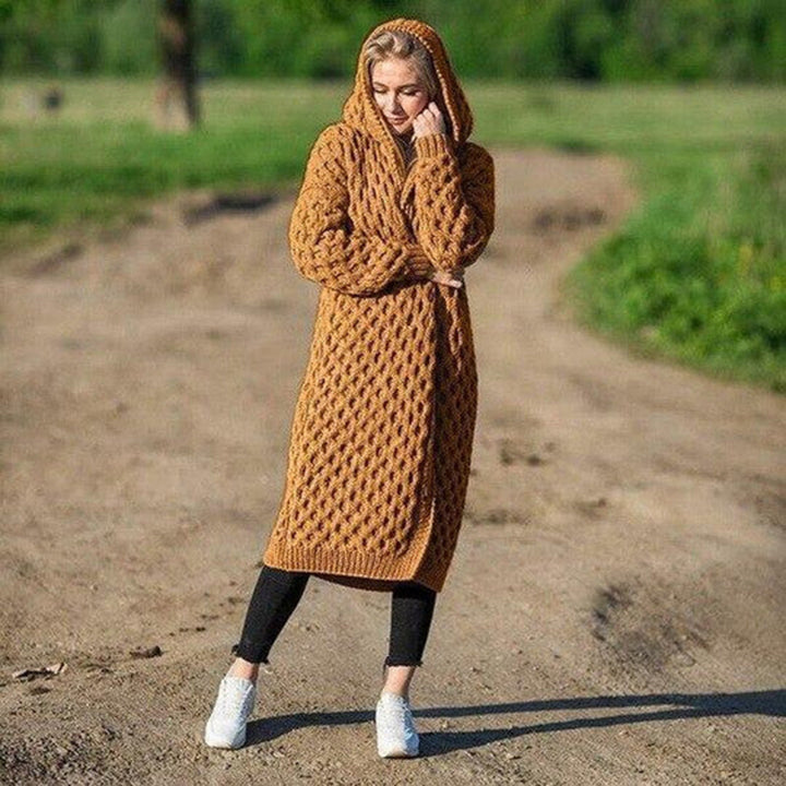 Frau in braunem Strickkleid mit Kapuze steht auf einem Feldweg, umgeben von grüner Natur. Modetrend, Strickmode, Herbstoutfit.
