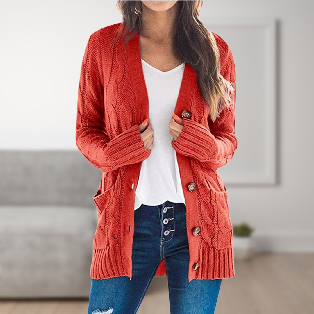 Frau trägt roten Strickcardigan mit Knöpfen, weißes T-Shirt und blaue Jeans. Modischer Herbst-Look, gemütlicher Wohnraum im Hintergrund.