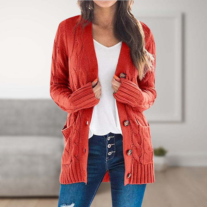 Frau trägt roten Strickcardigan mit Knöpfen, weißes T-Shirt und blaue Jeans. Modischer Herbst-Look, gemütlicher Wohnraum im Hintergrund.