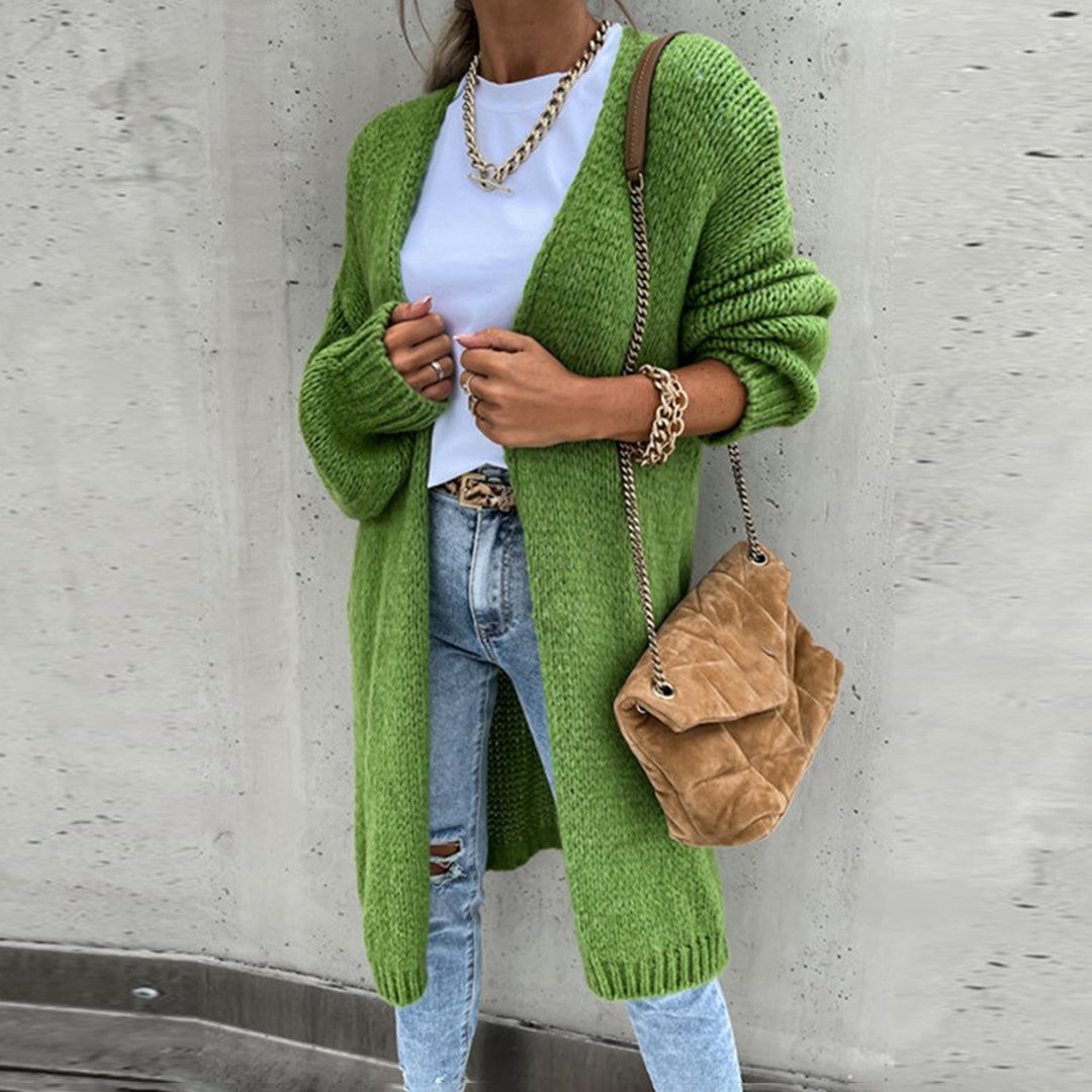 Frau in grünem Strickcardigan, weißem T-Shirt und blauer Jeans, trägt braune Umhängetasche und goldene Kette. Modischer Herbst-Look.