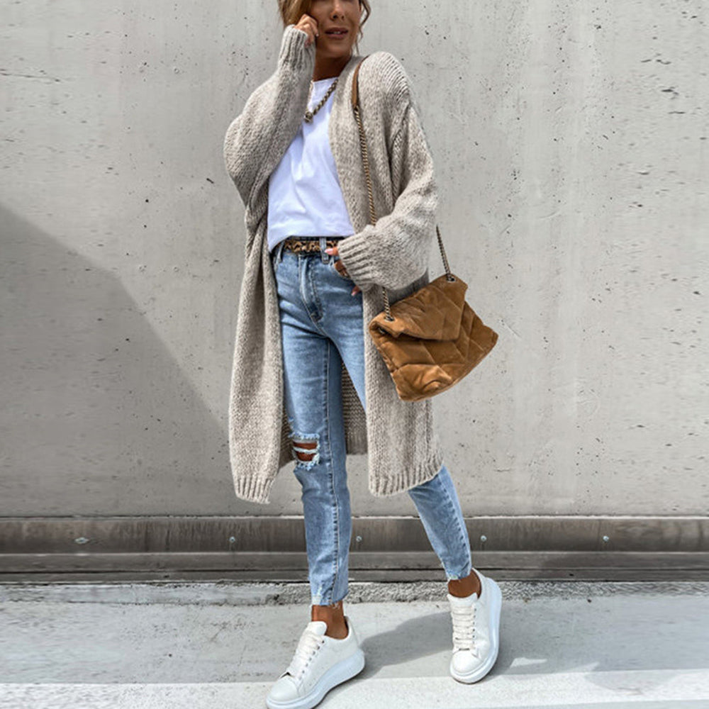 Frau in lässigem Herbstoutfit mit langem Strickcardigan, zerrissenen Jeans und weißen Sneakers vor Betonwand. Modetrend, Streetstyle, Damenmode.