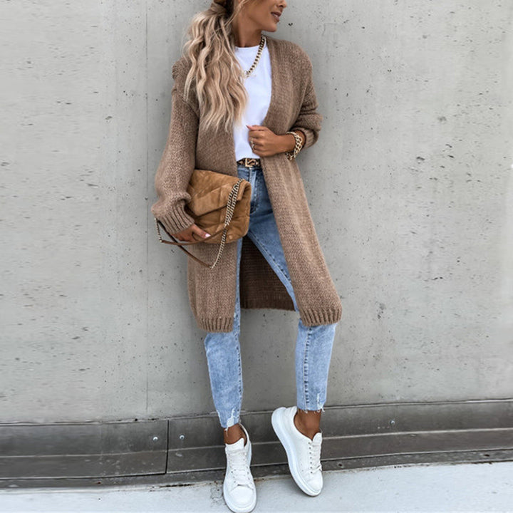 Frau in lässigem Herbstoutfit mit langem Strickcardigan, blauer Jeans und weißen Sneakers vor Betonwand. Trendige Mode, Streetstyle, Damenbekleidung.