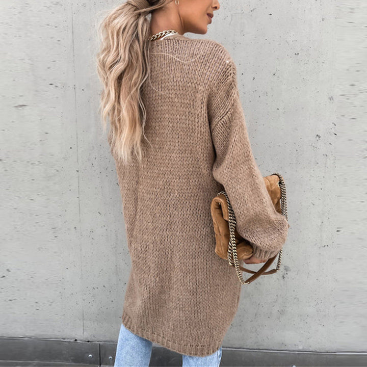 Frau mit langen blonden Haaren trägt einen beigen Strickpullover und hält eine braune Tasche mit Kettendetails. Modetrend, Herbstmode, Streetstyle.
