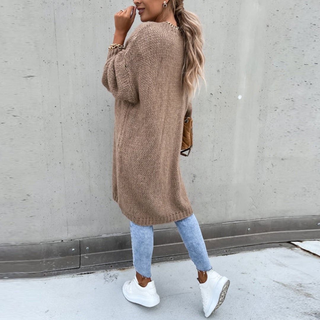 Frau in langem, braunem Strickpullover, blauer Jeans und weißen Sneakers vor grauer Wand. Modetrend, Herbstoutfit, lässiger Stil.