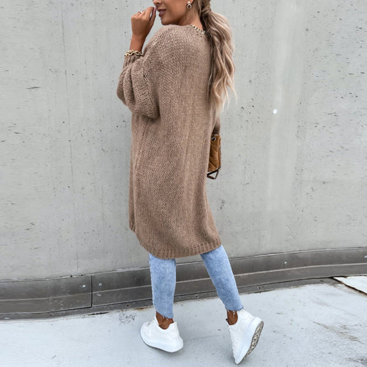 Frau in langem, braunem Strickpullover, blauer Jeans und weißen Sneakers vor grauer Wand. Modetrend, Herbstoutfit, lässiger Stil.