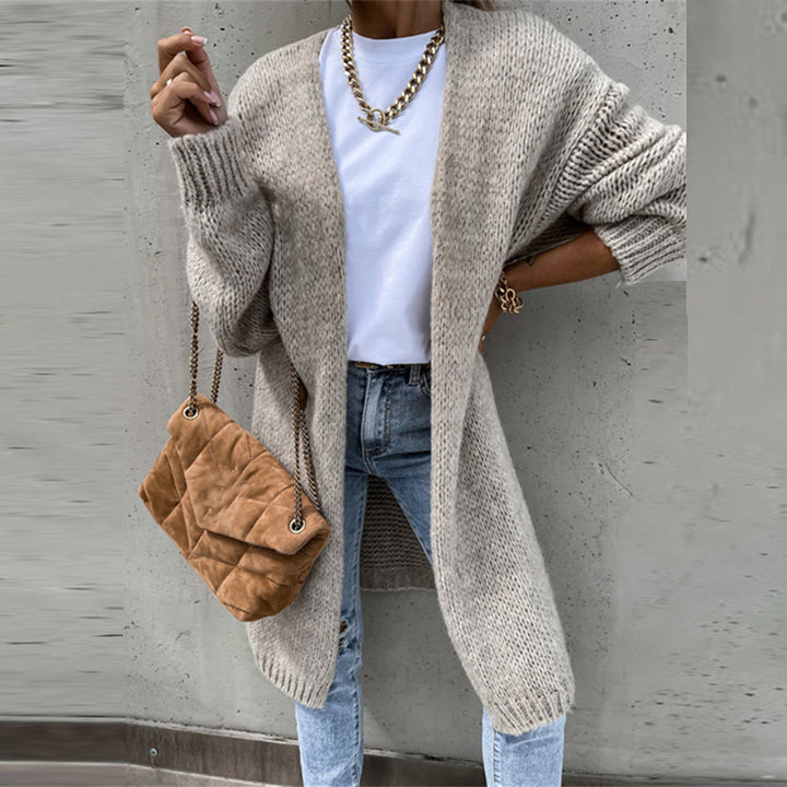 Frau in grauem Strickcardigan, weißem T-Shirt und blauer Jeans, trägt eine beige gesteppte Handtasche mit Kettengurt. Modischer Herbst-Look.