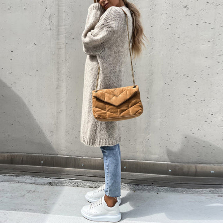 Frau in beigem Strickpullover, blauer Jeans und weißen Sneakers, trägt eine braune Umhängetasche. Urbaner Hintergrund, modischer Streetstyle-Look.
