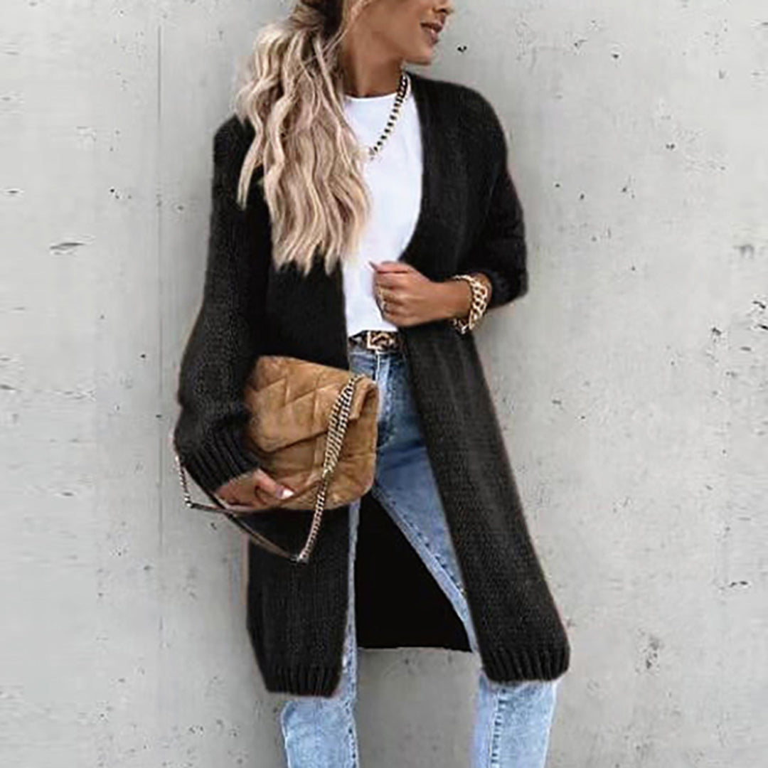 Frau mit langem, schwarzem Strickcardigan, weißem T-Shirt und blauer Jeans, hält eine beige gesteppte Handtasche, lässiger Herbstlook.