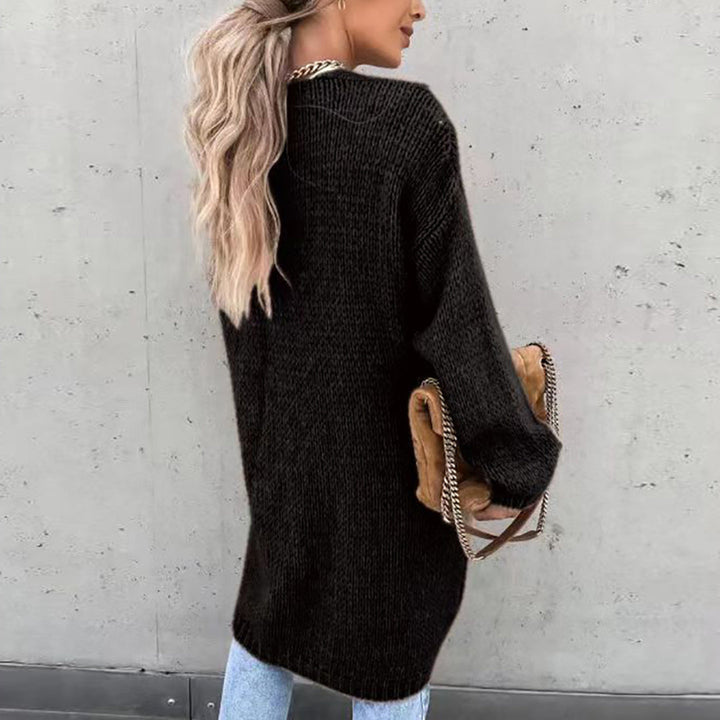Frau mit langen blonden Haaren trägt schwarzen Strickpullover und blaue Jeans, hält braune Tasche mit Kettendetails, steht vor grauer Wand.