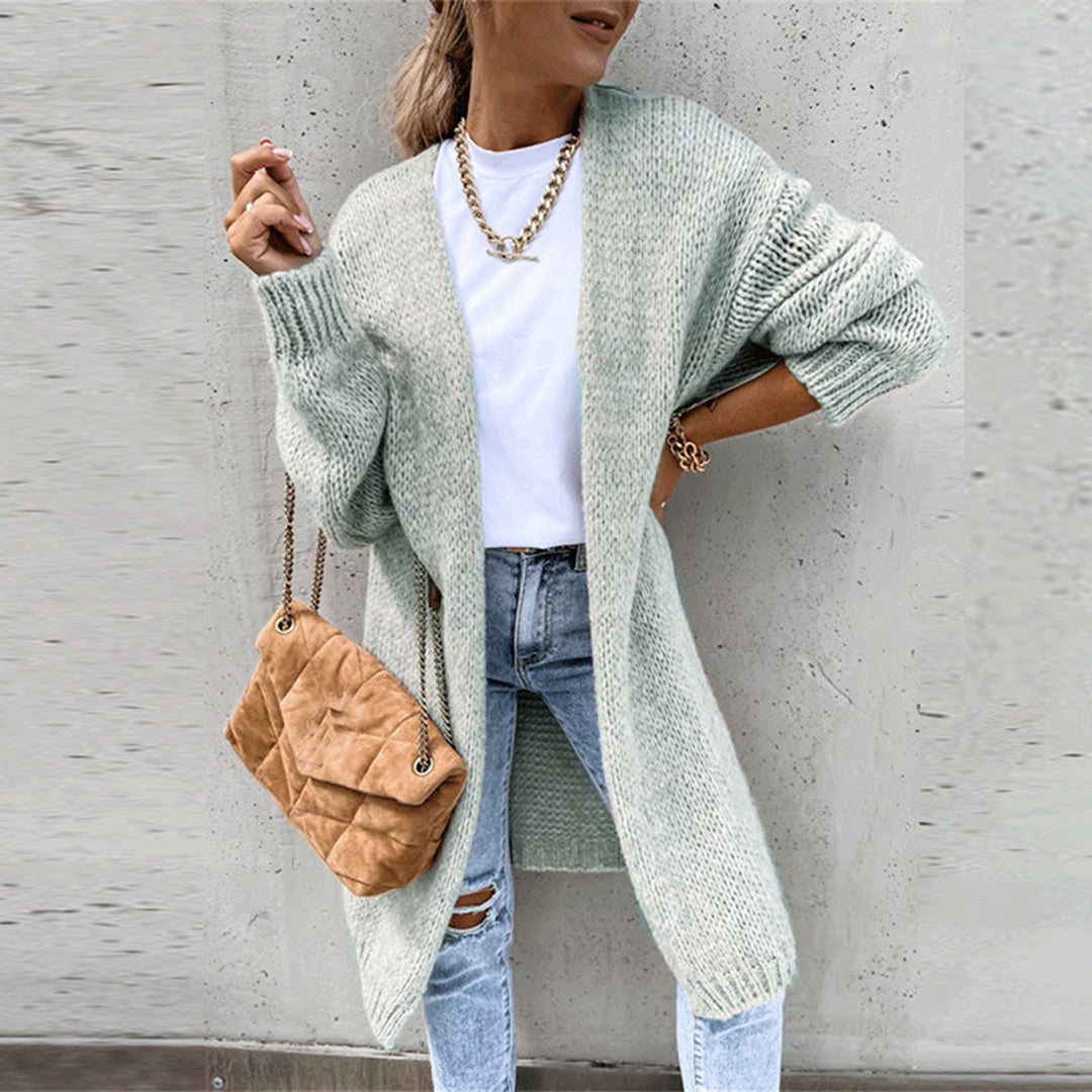 Frau in hellgrünem Strickcardigan, weißem T-Shirt und zerrissenen Jeans, trägt braune Umhängetasche und goldene Kette, vor grauer Wand.