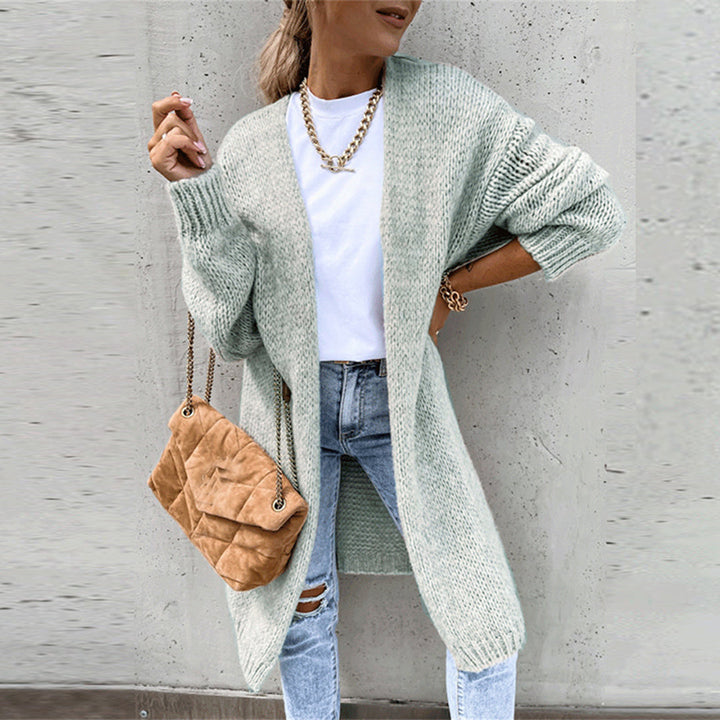 Frau in hellgrünem Strickcardigan, weißem T-Shirt und zerrissenen Jeans, trägt braune Umhängetasche und goldene Kette, vor grauer Wand.