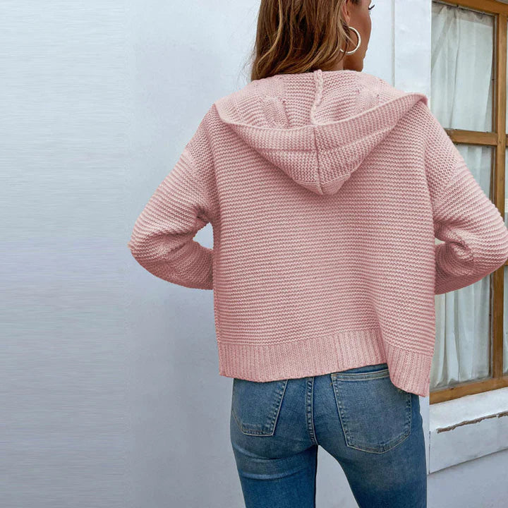 Frau in rosa Strickpullover mit Kapuze, Rückansicht, trägt blaue Jeans, steht vor einem Fenster. Modetrend, Damenbekleidung, lässiger Stil.
