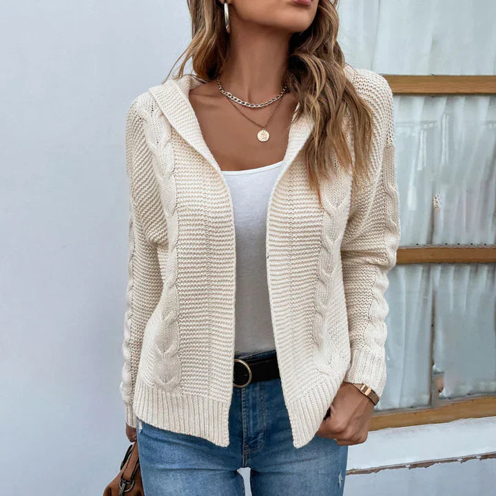 Frau in cremefarbener Strickjacke mit Zopfmuster, weißem Top und blauer Jeans vor einem Fenster. Modetrend, Damenmode, lässiger Herbstlook.
