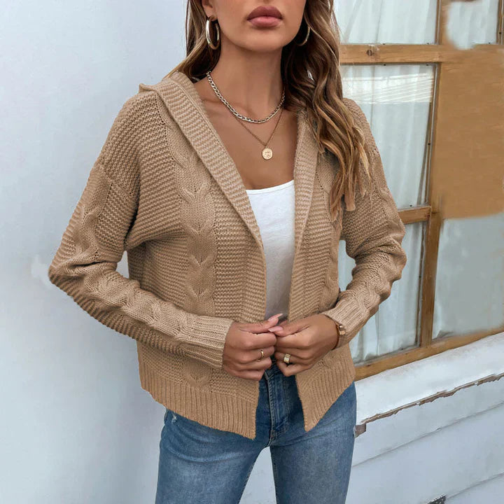 Frau trägt beige Strickjacke mit Zopfmuster, weißes Top und Jeans vor Fenster. Modetrend, Herbstmode, lässiger Stil, Damenbekleidung.