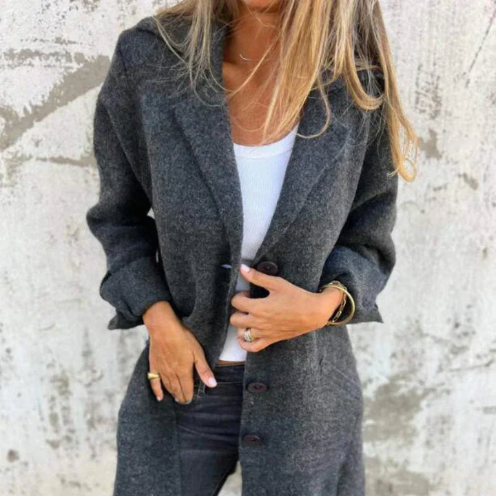 Frau in grauem Wollmantel, weißem Top und schwarzer Hose vor Betonwand. Modetrend Herbst, stilvolle Damenbekleidung, lässiger Look.