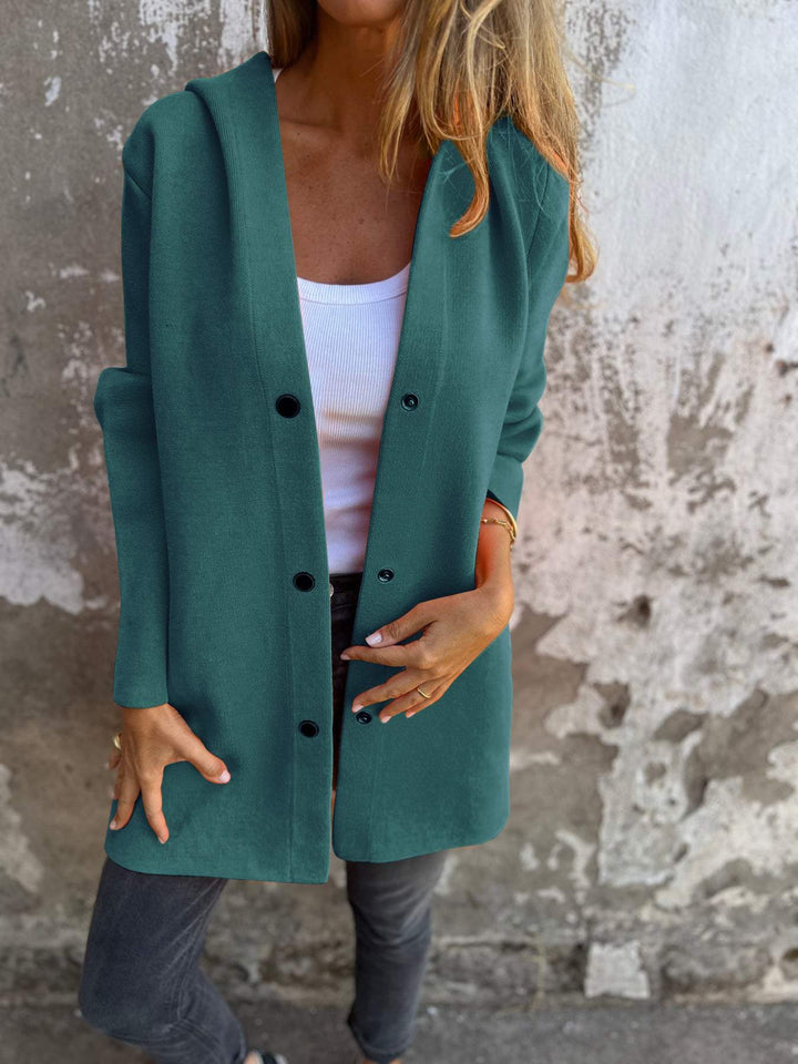 Frau in grünem Mantel, weißem Top und grauer Jeans vor einer verwitterten Wand. Modetrend, Herbstmode, lässiger Stil, Damenbekleidung.