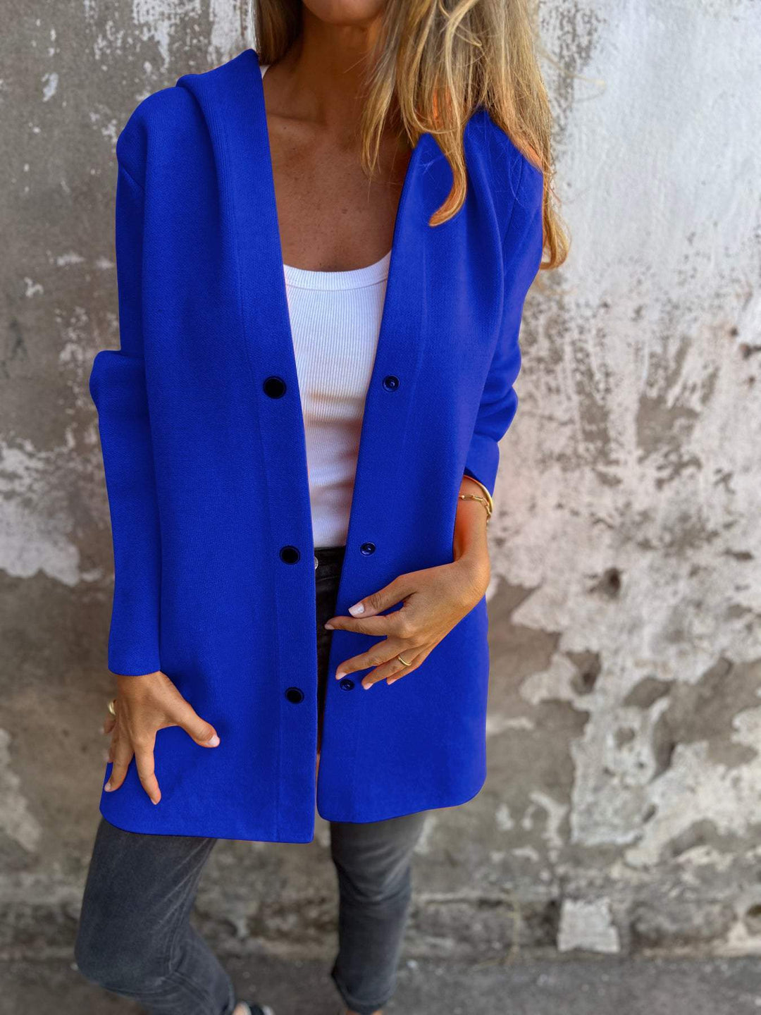 Frau in blauer Strickjacke, weißem Top und grauer Jeans vor einer Betonwand. Modetrend, Herbstmode, lässiger Stil, Damenbekleidung.