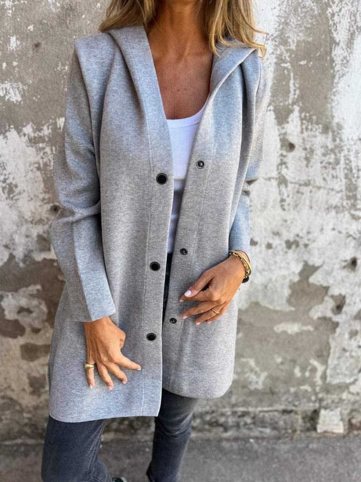 Grauer Damenmantel mit Kapuze, Knopfleiste und Taschen, getragen von Frau vor Betonwand. Modischer Herbstlook, lässig und elegant.