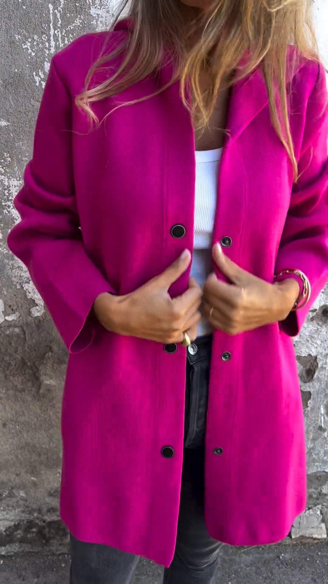 Frau in pinkem Mantel vor grauer Wand, trägt weißes Top und schwarze Hose. Modetrend, Herbstmode, stilvolle Damenbekleidung.