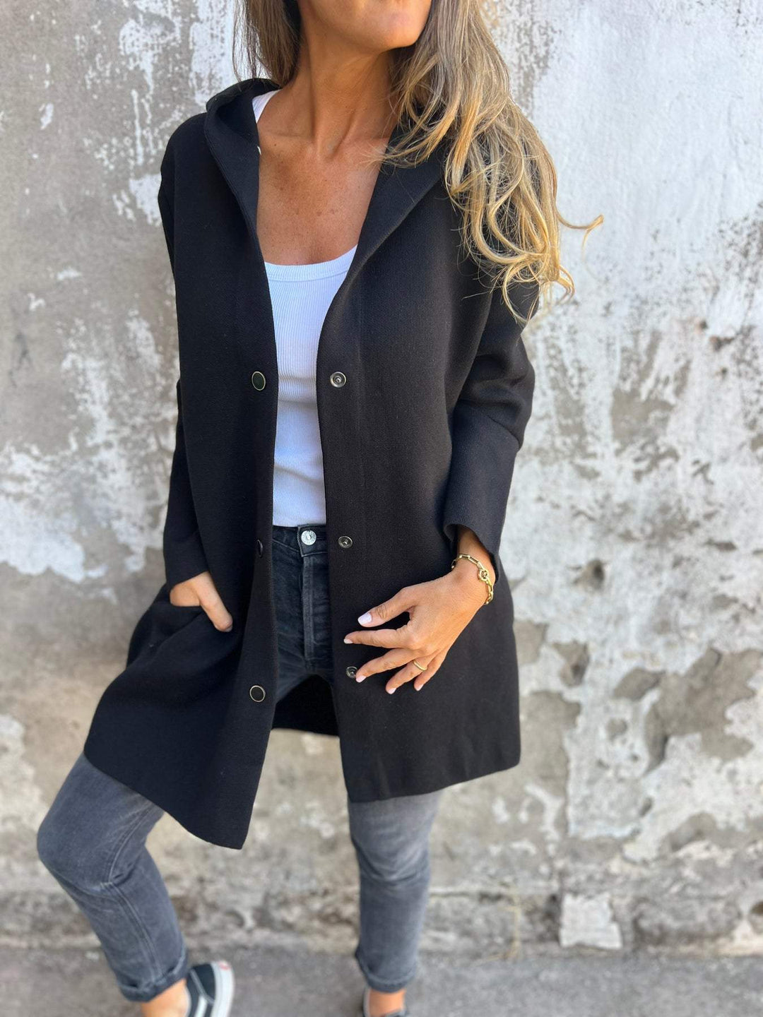 Frau in schwarzem Mantel, weißem Top und grauer Jeans vor einer Betonwand. Modischer Herbstlook, lässiger Stil, Streetwear.