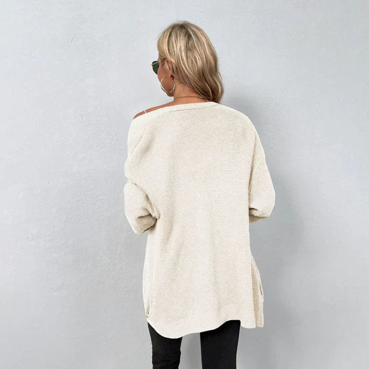 Frau mit blonden Haaren trägt einen cremefarbenen Oversize-Pullover und schwarze Hose, steht vor grauer Wand. Modetrend, lässiger Stil, Herbstmode.