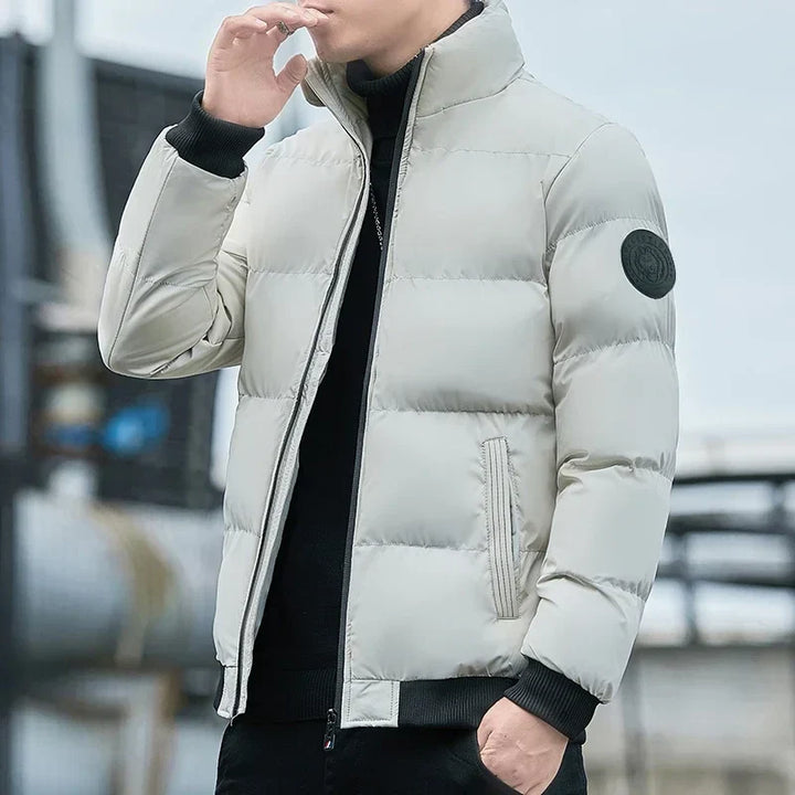 Mann in weißer Daunenjacke mit schwarzem Emblem, stehend im Freien. Modische Winterbekleidung, stilvoll und warm. Ideal für kalte Tage.