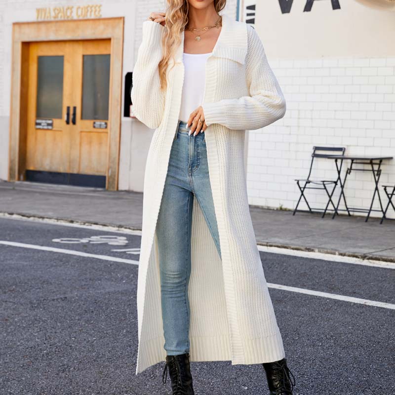 Frau in weißem Strickmantel und blauer Jeans auf städtischer Straße, modisch gekleidet, vor Café mit Holztüren. Trendige Herbstmode, lässiger Stil.