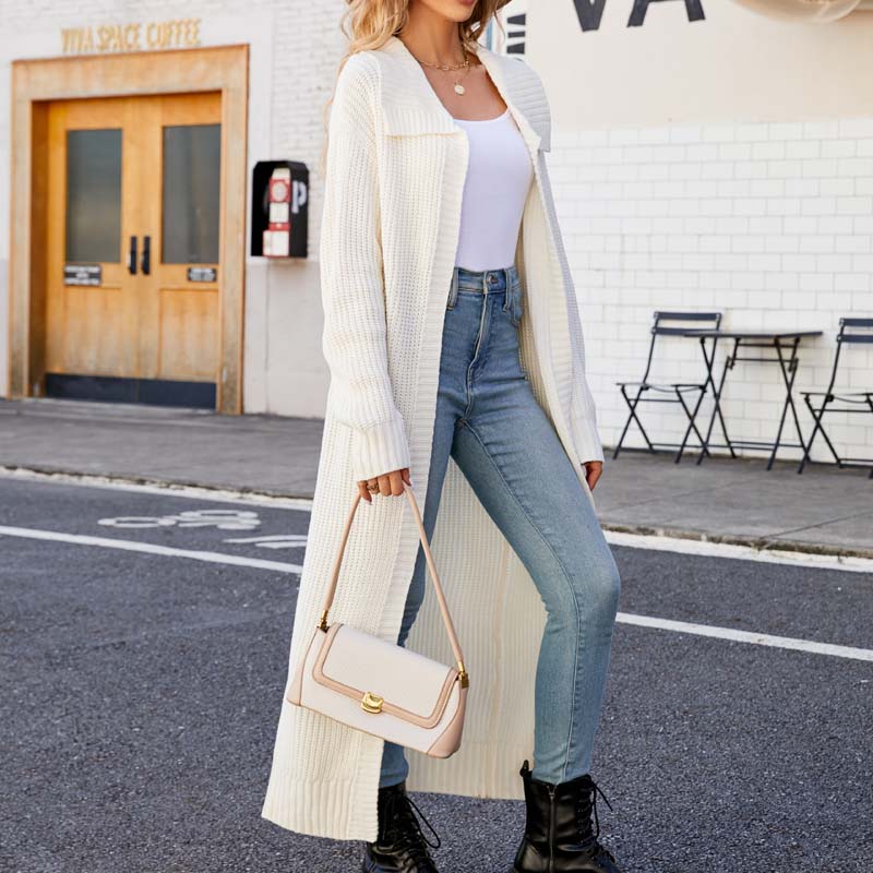 Frau in weißem Strickmantel und blauer Jeans, hält eine beige Handtasche, steht vor einem Café. Modetrend Herbst, lässiger Streetstyle Look.