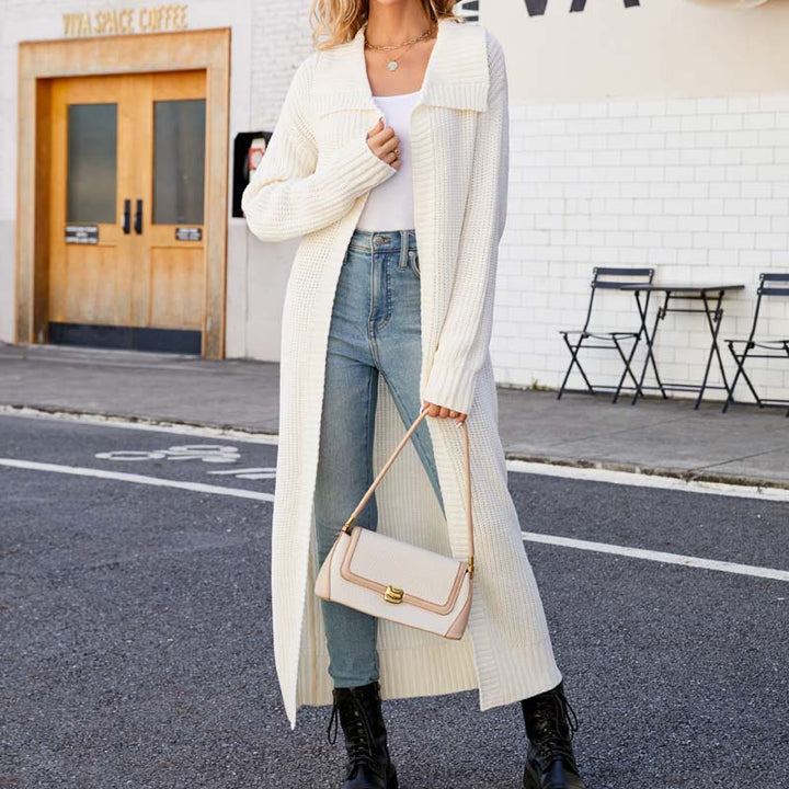 Frau in weißem Strick-Cardigan, blauer Jeans und schwarzen Stiefeln, hält eine beige Handtasche. Straßenhintergrund mit Café und Stühlen.