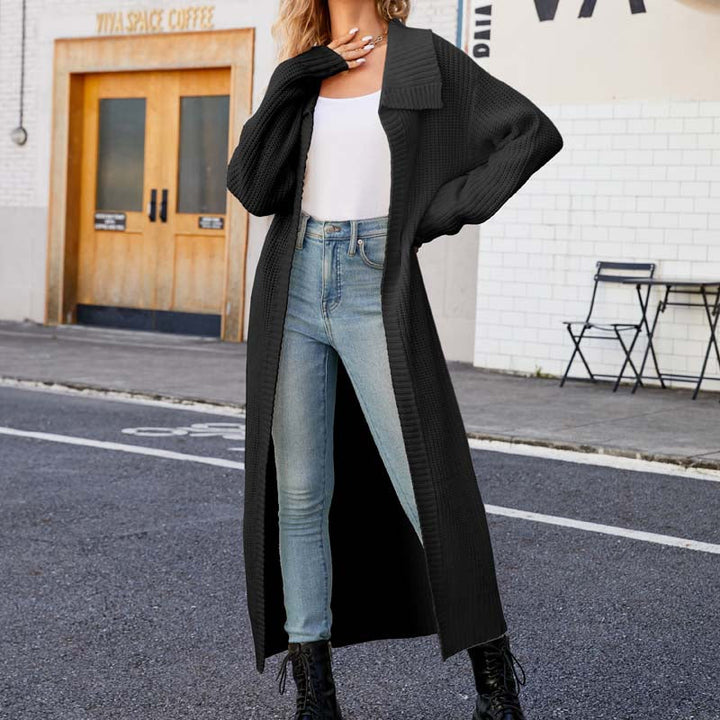 Frau in schwarzem Strickmantel, weißem Top und blauen Jeans posiert auf Straße vor Café. Modetrend, Herbstmode, lässiger Stil.