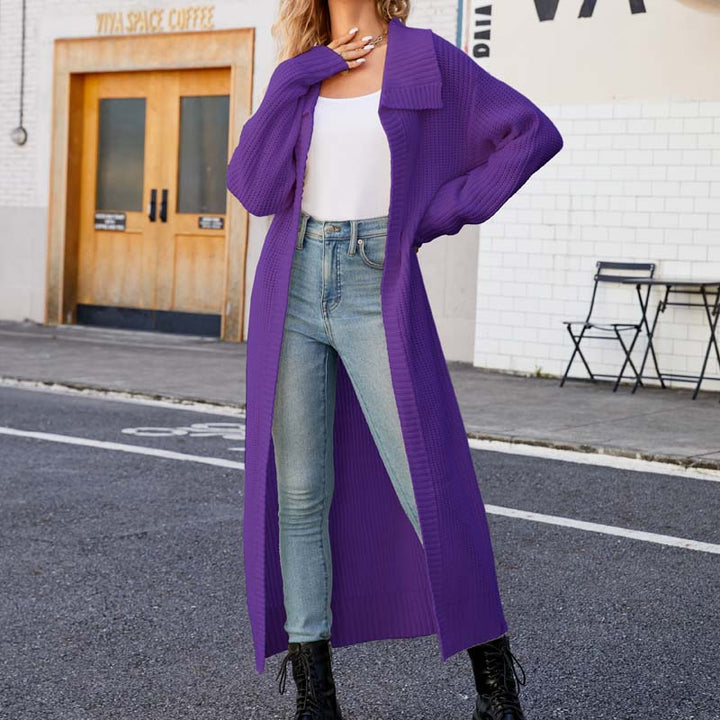 Frau in lila Strickmantel, weißem Top und Jeans posiert auf Straße vor Café. Modetrend, Herbstmode, lässiger Stil, urbane Kulisse.