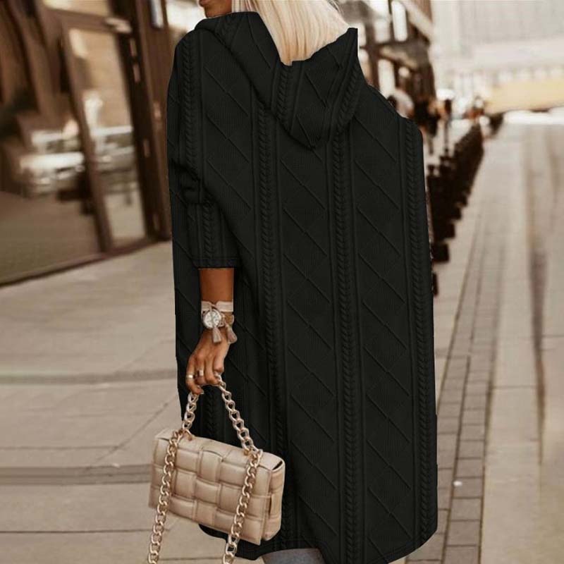 Frau in schwarzem Strickmantel mit Kapuze, hält beige gesteppte Handtasche mit Kettengriff, geht auf städtischem Gehweg. Mode, Herbst, Streetstyle.