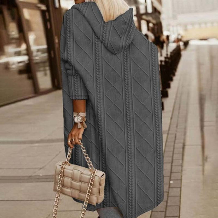 Frau in grauem Strickmantel mit Kapuze, hält beige gesteppte Handtasche, geht auf städtischem Gehweg. Modetrend Herbst, stilvolle Damenbekleidung.