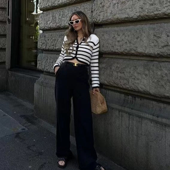 Frau in gestreiftem Pullover und schwarzer Hose, trägt weiße Sonnenbrille und Strohtasche, lehnt an einer Steinwand in der Stadt.