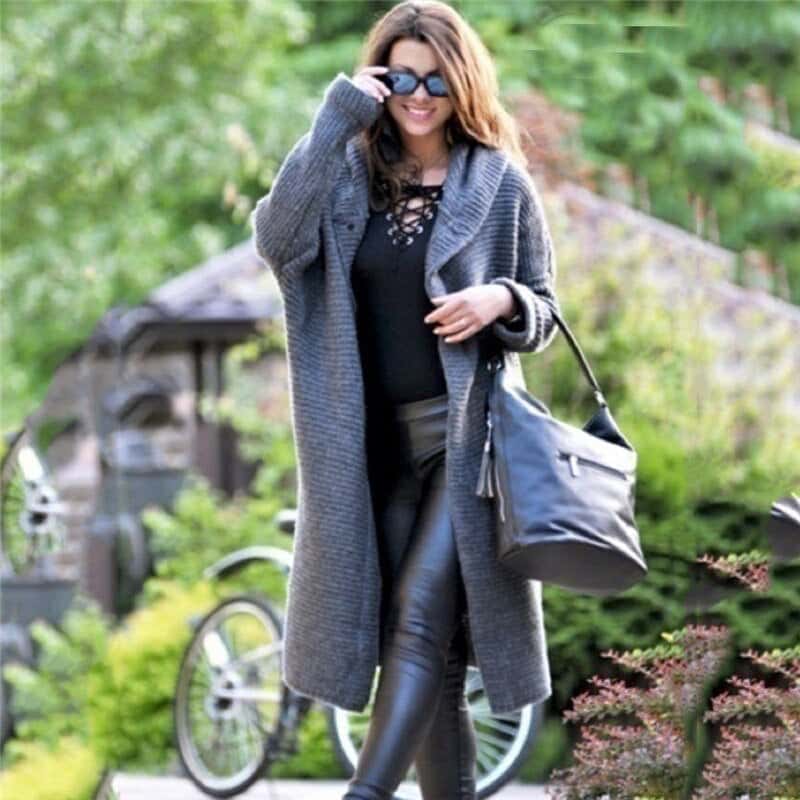 Frau in grauem Strickmantel und schwarzer Lederhose, trägt schwarze Tasche, steht vor Fahrrad im Grünen. Modetrend Herbst, lässiger Stil.