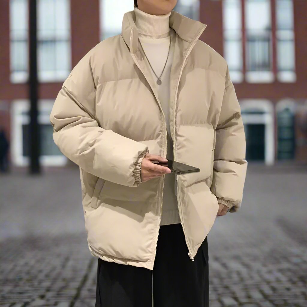 Person in beiger Daunenjacke mit Smartphone, steht auf gepflastertem Platz vor unscharfem Gebäude. Wintermode, Outdoor, urbane Umgebung.